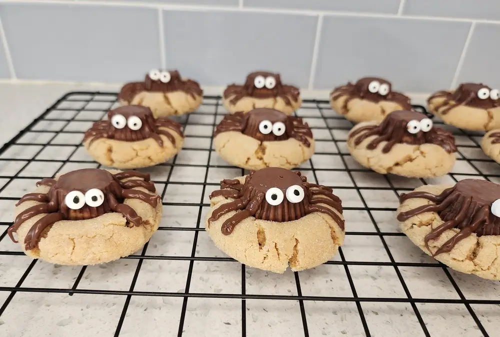 Homemade spider cookies with peanut butter bases, Reese’s cups, and candy eyes cooling on a wire rack after baking.
