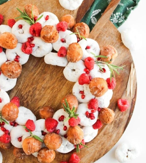 Mini donut Christmas wreath made with powdered donuts, donut holes, raspberries, pomegranate seeds, and rosemary, an easy no-bake holiday snack craft for kids.
