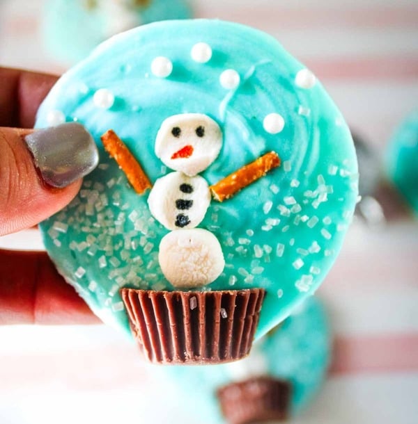 Snow globe cookies made with melted white chocolate, marshmallows, pretzels, and peanut butter cups decorated like snowmen, an easy Christmas treat for kids.