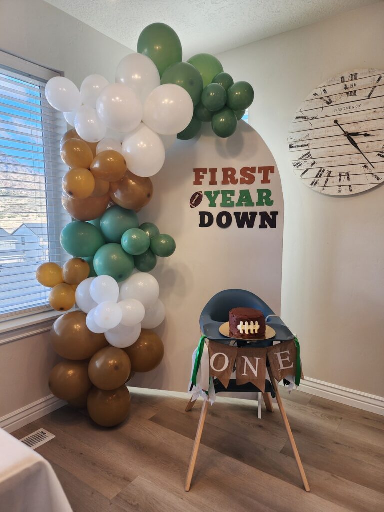 White arch backdrop with a green, brown, and white balloon arch, featuring a football-themed smash cake set up in front on a highchair